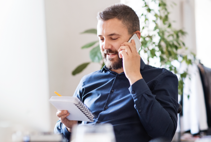 A person sits in a chair talking on the phone while looking at a notebook they are holding.