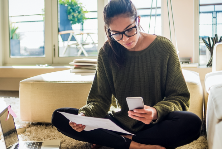 A person sits next to a laptop, looking at a phone screen in one hand while holding a piece of paper in their other hand. 