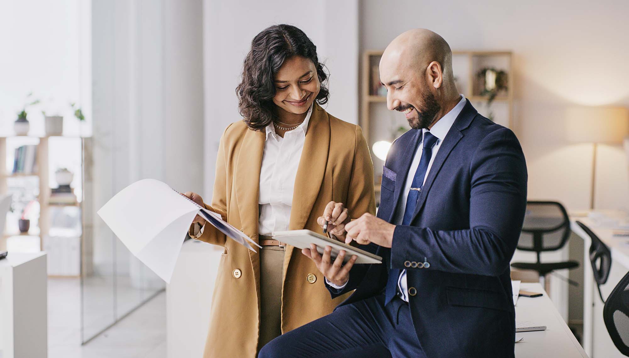 Two professionals smiling and working together, one writing notes and the other using a tablet.